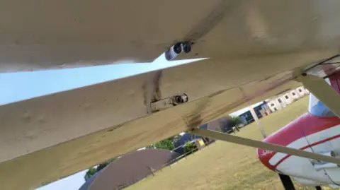 A close-up picture of the wing of a plane where a pin is missing. The plane is white and the nose of it is red.