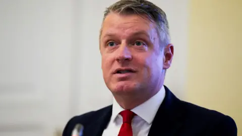 Luke Pollard, a man with grey hair in a dark suit, white shirt and red tie stands in front of a small lectern microphone.