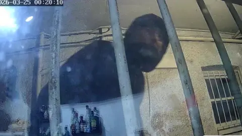 A man dressed in black, with a hood, seen through a widow with bars