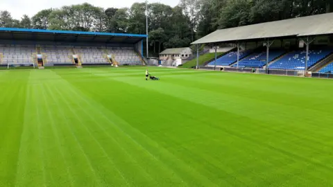 Local Democracy Reporting Service A large, well-maintained football stadium with a freshly cut green pitch. In the centre of the field, a single groundskeeper is pushing a lawn mower, creating neat, light and dark striped patterns across the grass. The pitch stretches widely across the foreground, appearing smooth and evenly trimmed.
On the left side of the image, there is a covered stand with rows of empty seating arranged in tiers. The structure has a blue roof and metal framework, with stairways painted in yellow leading up through the seating area. A small football goal is positioned near this stand at the edge of the field.
On the right side, there is another stand with blue seats arranged in orderly rows under a sloped roof. This stand appears slightly larger and sits on a raised grassy embankment. Behind this stand and between the two seating areas, a small building with a light-coloured exterior is visible, along with a fence and some basic stadium infrastructure.
In the background, a dense line of tall green trees surrounds the stadium, forming a natural backdrop. A tall floodlight pole rises above the trees near the centre of the image.