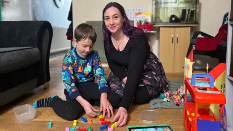 Jace wearing a blue sweatshirt with colourful graphics and black trousers sitting next to Sarah wearing a black shirt and a black and pink floral skirt playing with logos