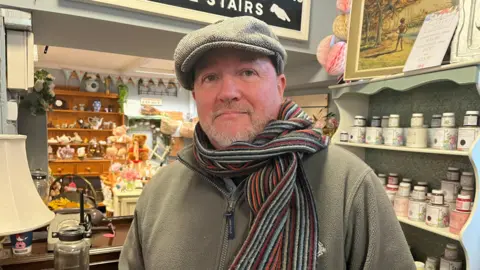Allun is standing inside a vintage-style shop filled with decorative items. The setting includes shelves lined with jars, a display of teacups and saucers, and a wooden counter with a lamp and glassware. He is wearing a grey fleece jacket, a flat cap, and a long striped scarf draped around the neck.