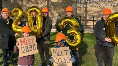 Jo Rust Four people standing outside the Queen Elizabeth Hospital in King's Lynn. They are wearing orange hard hats. Each protester is holding a large gold balloon. The balloons spell out 2030.