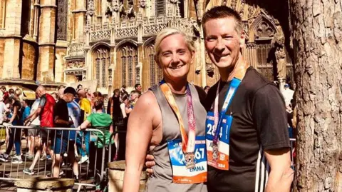 Submitted A man with short brown hair and a black top is wearing a running sticker on his top and a medal around his neck. He has his arms around a woman with blonde hair tied back who is wearing a medal round her neck and a running sticker on her brown vest. Behind them you can see metal railings and several people dotted about next to part of a cathedral.