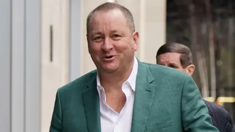 Mike Ashley smiling wearing an open-collar white shirt and a green suit jacket
