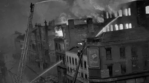 David Facey Bristol Archives A Filton building which had just been bombed. The photo is in black and white. Thick clouds of smoke are billowing from the roof and the windows are lit up with the light of the blaze. Men with white helmets are stood on ladders that reach the height of the building with large hoses sending streams of water across the roof and through windows.
