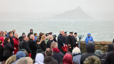 Penzance Council A crowd at a remembrance service in Penzance. The crowd are smartly dressed. A stormy sea is behind the sea wall. Fog is blurring the view in the distance.