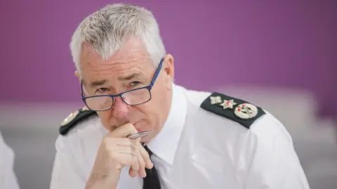 PSNI chief constable Jon Boutcher, a man with grey hair, wearing a white shirt and black tie. He is also wearing glasses and has his hand to his mouth. 