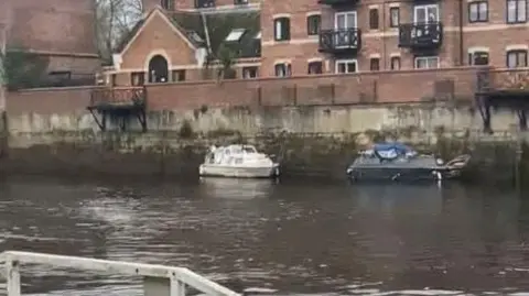 Will Tretheway A calm riverside scene featuring small docked boats and a large brick residential building with balconies and varied windows. 