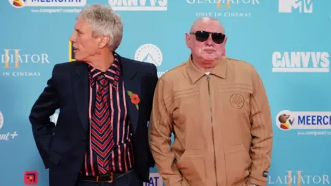 PA Media Two men pose for the cameras at a press event. One is wearing a beige jacket and sunglasses, the other a suit jacket with striped shirt and tie