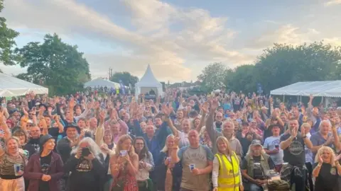 A large crown of 100s of people look towards the camera dressed in an array of colours. Some of them hold their hands in the air. The are all outside and the sky is blue with white clouds. In the distance there are several tents and gazebos.