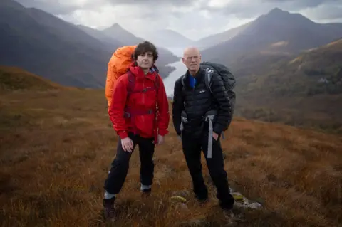 Rob Leech Danny and Phil are wearing outdoor clothing. They are carrying rucksacks. The men are standing on a hill with loch and mountains behind them.