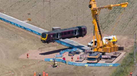 A giant yellow crane in a field next to railway line. It is lifting a train carriage. A temporary track has been built to get the crane into position in the field. There are about a dozen people in orange overalls.