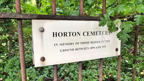 a plaque fixed to a metal railing, inscribed with the words "HORTON CEMETERY in memory of those buried in this ground between 1899 and 1955"