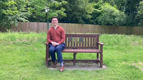 Kate Moser Andon/BBC Luke sitting on a bench on a green in Fulbourn