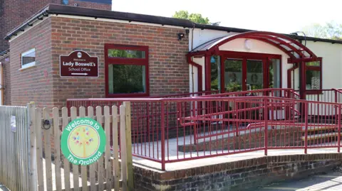 The outside of a primary school. It is a single storey building with red railings and a ramp leading to the main door. The building is brown brick on the left and white on the right