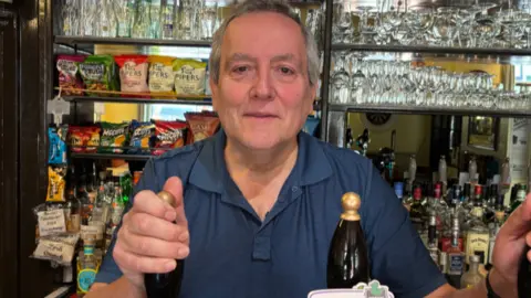 John is stood behind the bar with beer pumps in front of him and rows of alcohol behind him

