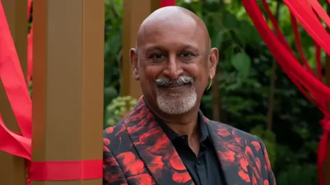 Tackle HIV Manoj Malde surrounded by the red ribbons attached to the seating area. Th red ribbons are a symbol of HIV awareness and support. He is wearing a red and black suit, has a gold hopped earrings in his right ear and a grey moustache and goatee.