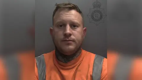 Police custody photograph of Ben Whitby. He has short dark hair and beard, tattoos around his neck and is wearing an orange high-visibility top