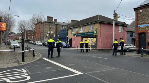 Six police officers wearing yellow and black uniforms stand in the street with a police cordon. The street is lined with cars, shops and houses. The sky is cloudy and grey. 