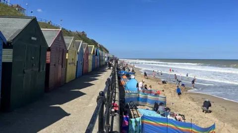 Andrew Turner/BBC Mundesley seafront with beach huts and bathers on beach