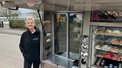 A woman with blonde hair, wearing a black hooded top, is stood alongside the back of a van, which has a range of food on display on metal shelves