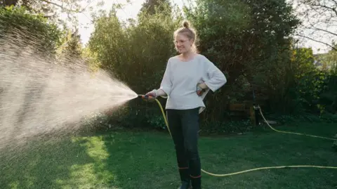 Getty Images A woman is watering the lawn in a garden with a hosepipe. The water is lit up by sunshine. She has her hair in a bun and is wearing glasses. The sunlight is filtering through the shrubs and trees.
