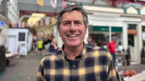 A middle-aged white man smiling at the camera. He has dark grey hair and is wearing a yellow and black checked shirt.