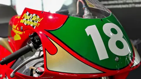 The bike raced in the lightweight 250 race that year, it is red with yellow logos and a green plate with the number 18 on the front.
