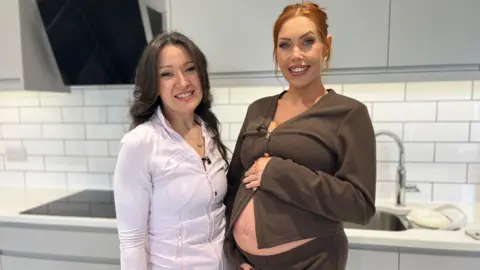 Rhiannan and Hayley are standing in a white kitchen. Rhiannan has long dark wavy hair and is wearing a white zip-up top. Hayley is holding her baby bump. She has red hair tied up in a bun and is wearing a brown cardigan which is partially open revealing her baby bump. 