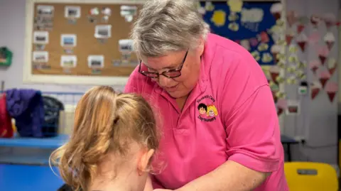 Shelia crouches down in front of a young girl who is facing away from the camera. They are inside a classroom. Shelia is helping her with some work. 