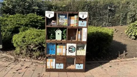 A bookcase promoting the Top of the Wold play standing on a path with greenery in the background, There is a bird box and and a number of brightly-coloured containers on it.