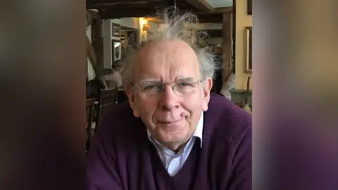 Family handout A picture of Léon Ellison, wearing a light shirt and a purple jumper, sat in a restaurant or pub. He has white hair and glasses.