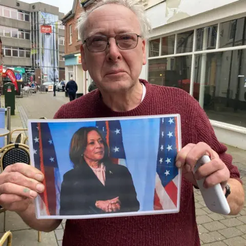 Julian Cunnington, with short grey, whispy hair, is holding a picture of Kamala Harris 