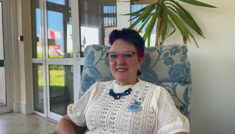 A woman with short, purple hair sat in a blue and white chair. She is wearing a white, short-sleeved knitted jumper with a blue necklace