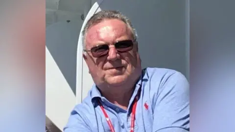 Wroxham Parish Council Peter Mantle who is wearing sunglasses, is looking directly at the camera and smiling. He is wearing a short sleeved blue T-shirt.