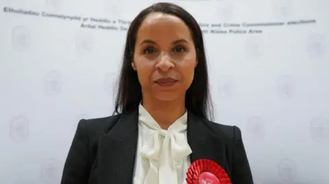 A head and shoulders picture of Emma Wools. She has black shoulder-length hair and is wearing a dark jacket with a red Labour rosette on it and a white blouse.