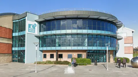 Google Redcar and Cleveland College is a three-storey, mainly glass building, with a curved exterior. A blue and white sign on the upper left of the complex reads: "Redcar and Cleveland College". There are two lampposts out front and people walking into the building.
