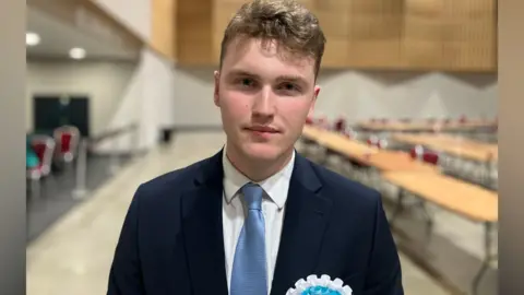 Sam Read/BBC Finlay Hughes looks at the camera wearing a dark blue suit jacket, pale blue tie and white shirt. The tables behind him are all empty.