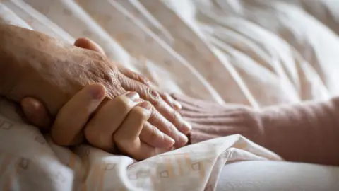 A hand hold's an elderly person's hand above a bed sheet