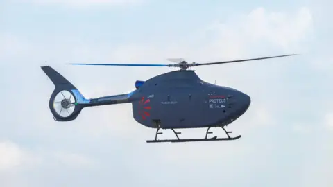 A navy blue helicopter. Its main rotor system is rotating. It is in the sky. The sky is blue with white clouds. 