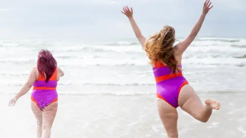 The Body Happy Organisation Two woman in pink and orange bathing suits run into waves on a beach.  They have their backs to the camera and water is splashing up their legs