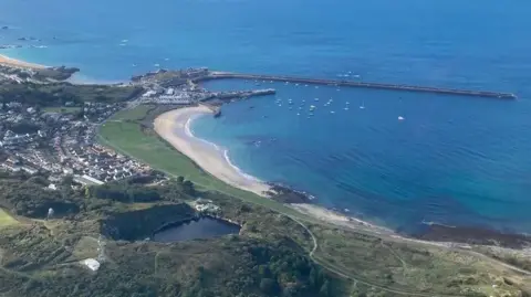 BBC A beach, bay and harbour in Alderney photographed from above on a clear day. There is a reservoir inland on the outskirts of a village. There are many yachts moored in the bay and a road runs along the coast.