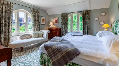Oliver's Travels Master bedroom which has a carpet patterned with green leaves, a large double bed, a chaise long and big windows with views onto the garden. 