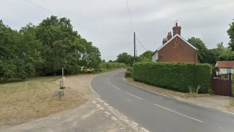 A street view image of Dedham Road. It is a country road with a house on one side and trees on the other.