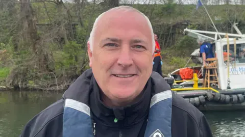 A man with short, white hair wearing a black coat and a blue and black life jacket.