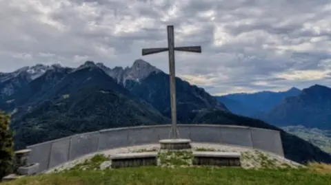 Submitted A large cross on top of a raised platform has a grey barrier in front of it. It overlooks several mountain tops on a cloudy day.