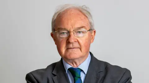 PA Media Declan Morgan is pictured against a grey background. He has short grey hair and is wearing silver rimmed glasses. He's dressed in a dark coloured suit, blue shirt and a blue and green tie. 