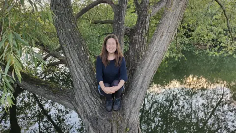 Bryony Parrish Bryony Parrish smiling for the camera while sitting in a tree. There is a body of water underneath.