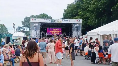A view of the main stage at the festival showing crowds of people standing and sitting around festival tents. 